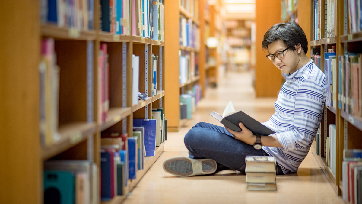 A student sitting on the floor of a library aisle reading a book