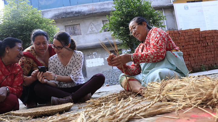 A group of women sitting outdoors weaving straw and looking at a smart phone