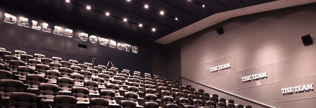 A large, modern theater-style room with "Dallas Cowboys" and "The Team" displayed on the walls. Rows of black leather chairs are arranged in a stadium-style layout under soft lighting.