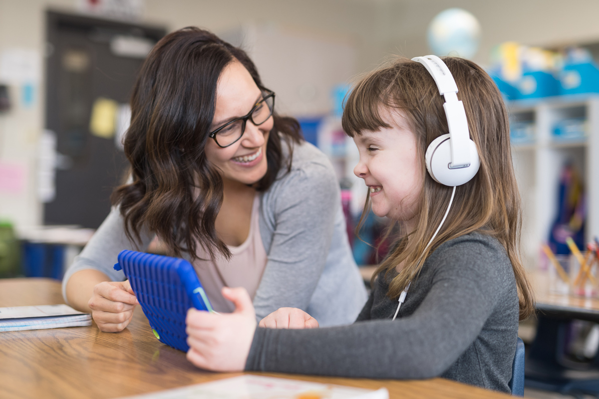 A smiling teacher sitting beside a girl with headphones and a  tablet