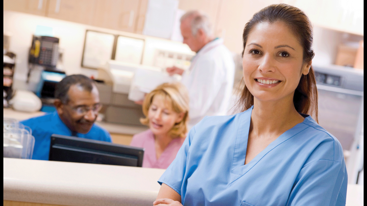 A smiling nurse at reception desk with colleagues working behind her