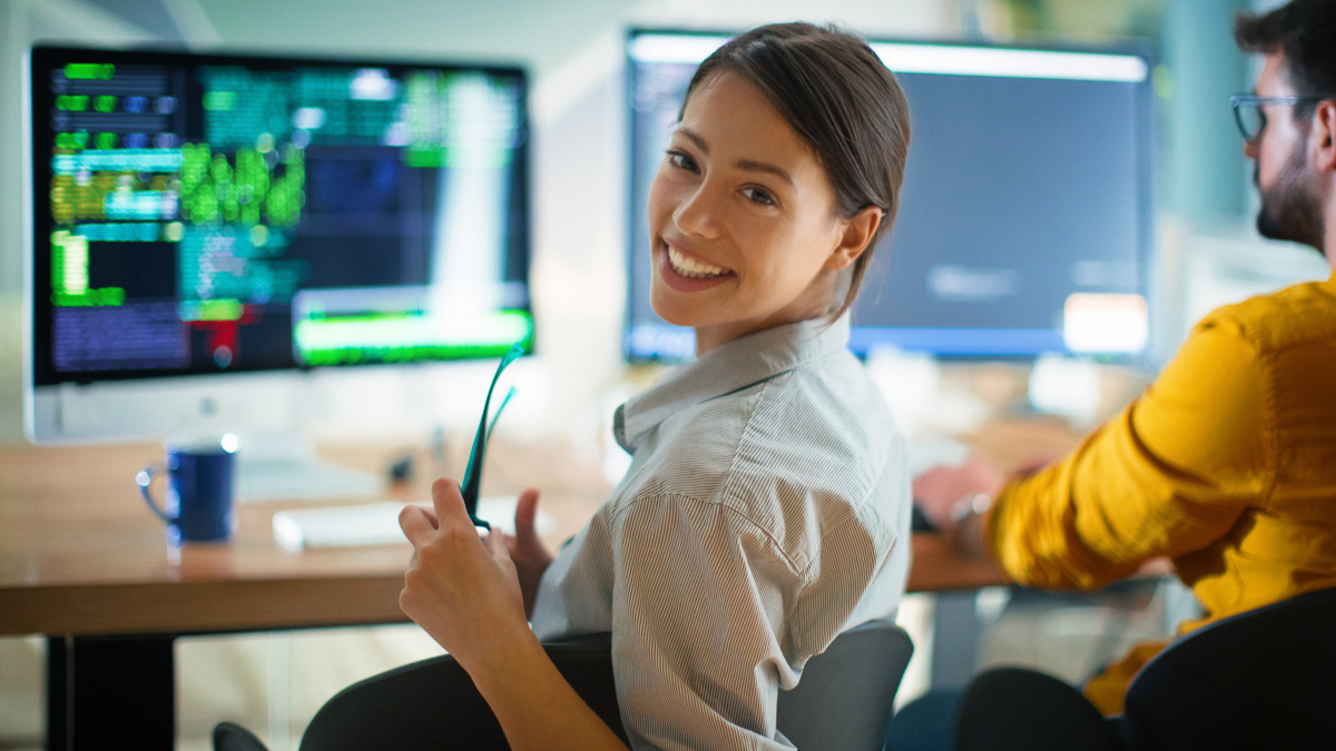 A woman smiling at her desk with code displayed on multiple monitors