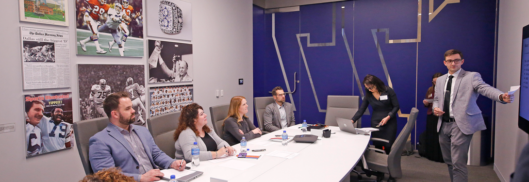 A professional meeting room with six people seated at a conference table and a presenter in a suit gesturing toward a screen. The table holds laptops, documents, and water bottles, with a sports-themed, memorabilia-decorated wall in the background.