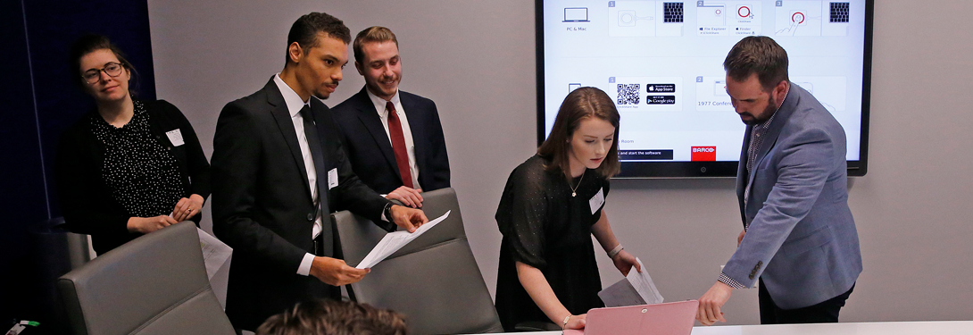 A group of professionals collaborating around a conference table, with one individual pointing at a laptop while others hold documents. A presentation screen displaying visuals is visible in the background.