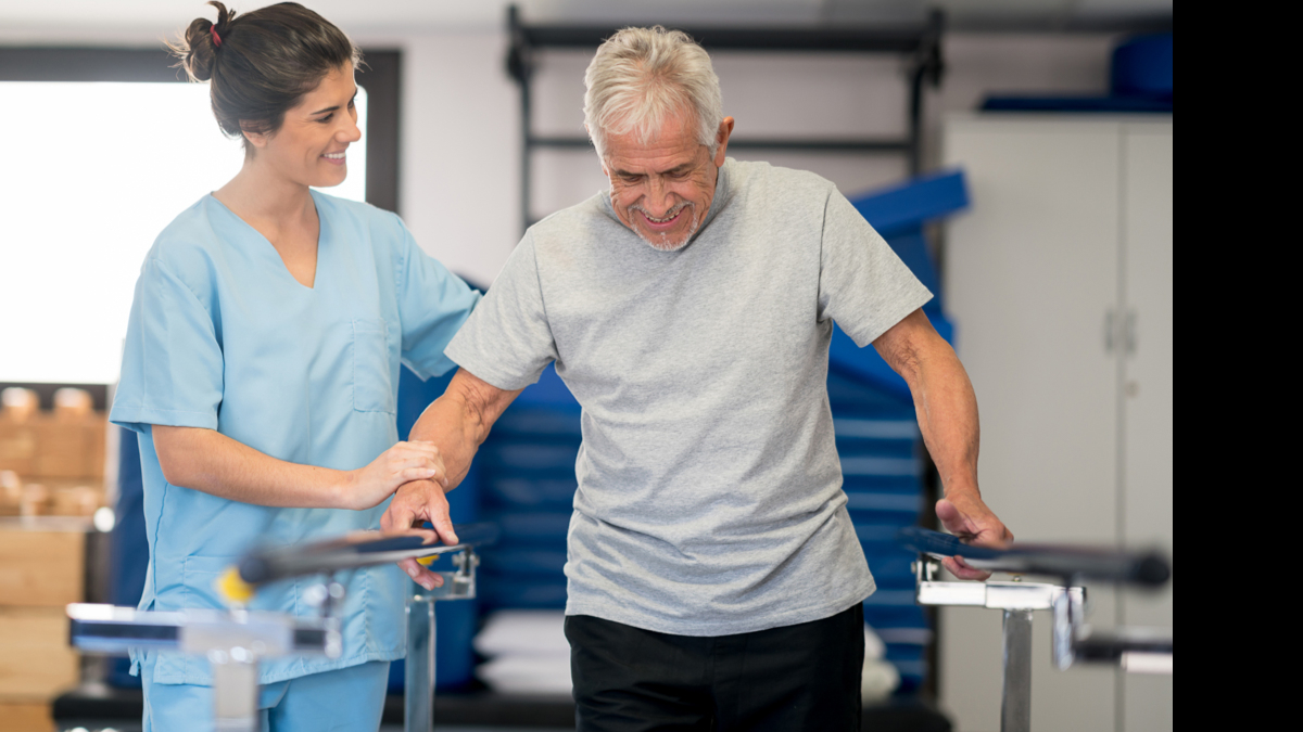 A physical therapist supporting an elderly man walking in a rehabilitation setting