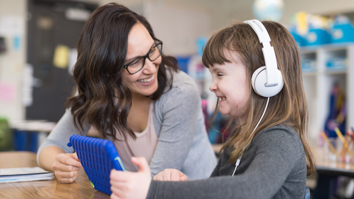 A smiling teacher sitting beside a girl with headphones and a  tablet