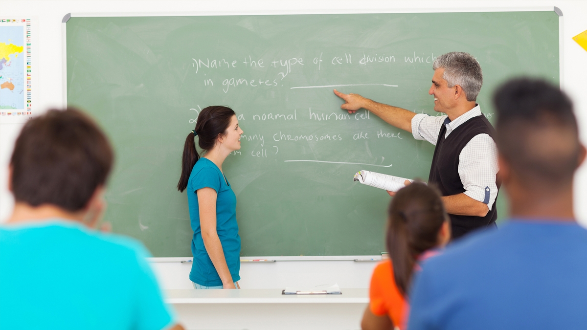 A science teacher at a chalkboard pointing out a question 